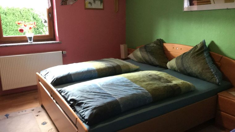 A cozy bedroom with a double bed and colorful pillows. The walls are painted in soft shades and there is a window overlooking green scenery.