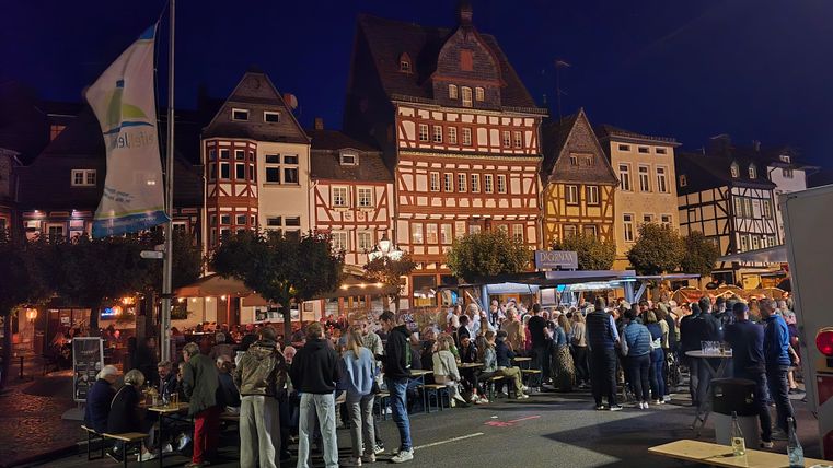 Une place de marché animée la nuit avec des maisons à colombages historiques. Des gens se rassemblent tandis que des stands proposent de la nourriture et des boissons.