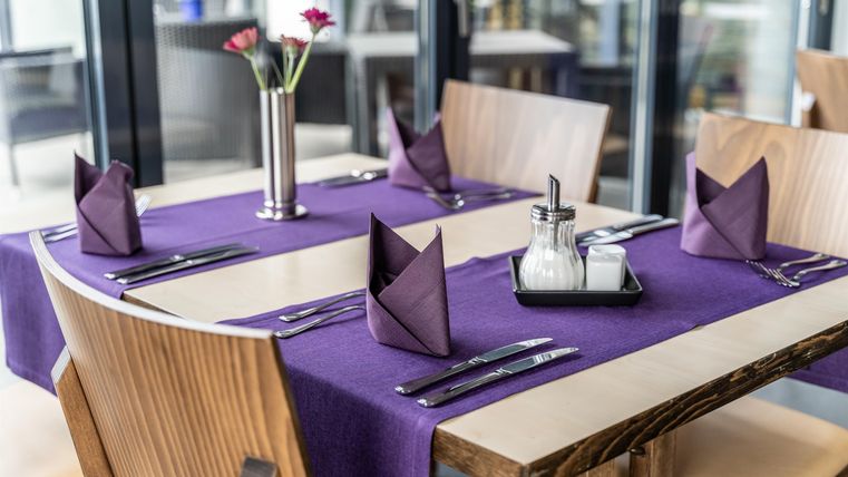 Een elegant gedekte tafel in een restaurant. De tafel heeft paarse tafelkleden, gevouwen servetten en een bloemenvaas in het midden.