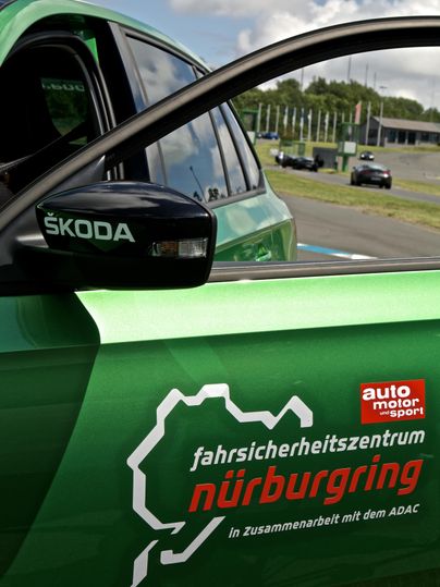 Ein grünes Auto am Nürburgring mit einem Aufkleber des Fahrsicherheitszentrums. Im Hintergrund sind Teile der Rennstrecke und weitere Fahrzeuge zu sehen.