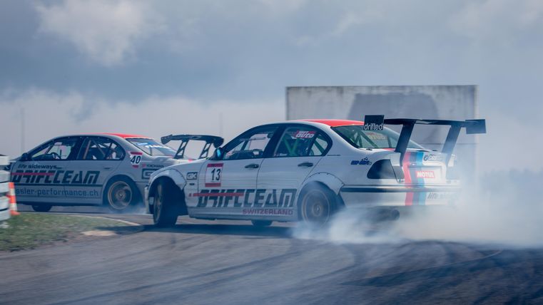 Zwei Rennwagen driften auf einer Piste und erzeugen großen Rauch. Der Himmel ist bewölkt und die Atmosphäre ist spannend.