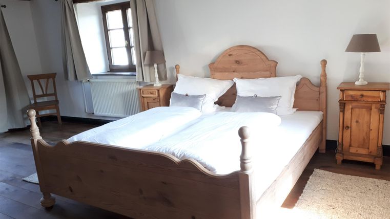 A cozy bedroom with a large double bed and a wooden frame. The room is bright and inviting, overlooking a small table and a lamp.