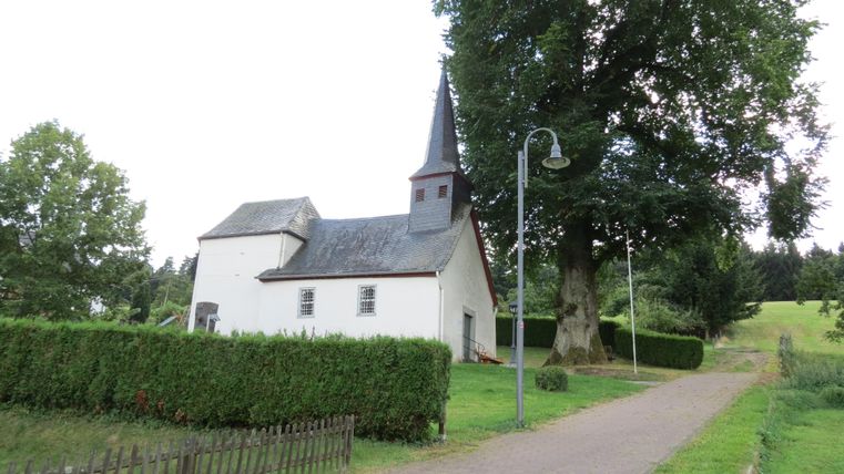 A picturesque church with a steep tower stands at the end of a path. Green trees and a hedge surround the building.