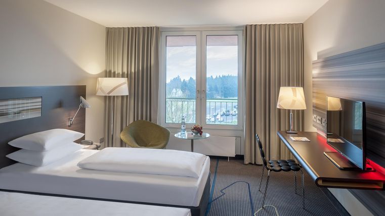 Een moderne hotelkamer met twee eenpersoonsbedden en een bureau. Grote ramen bieden uitzicht op de omgeving en veel natuurlijke verlichting.