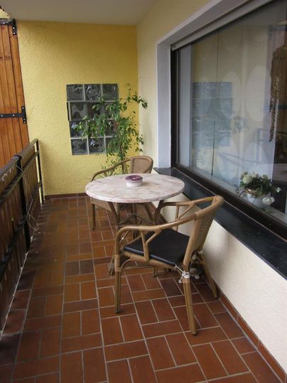 Een klein balkon met een ronde tafel en twee stoelen. Op de achtergrond staat een plant voor een raam.