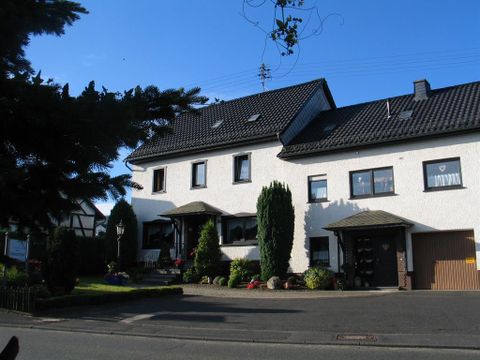 A beautiful two-story house with a well-maintained garden. The facade is painted white and there are some trees and plants around the building.