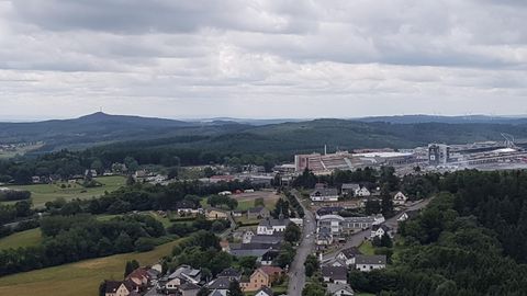 Eine Aussicht auf eine Stadt umgeben von grünen Hügeln und Wäldern. Im Hintergrund sind Gebäude und eine Gebirgskette zu sehen.