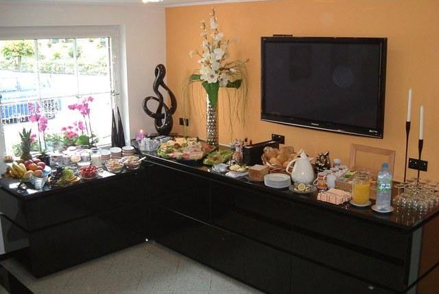 Ein gedeckter Tisch mit einer Vielzahl von Speisen und Getränken. Im Hintergrund sind Fenster mit Blick auf Pflanzen und eine moderne Einrichtung zu sehen.