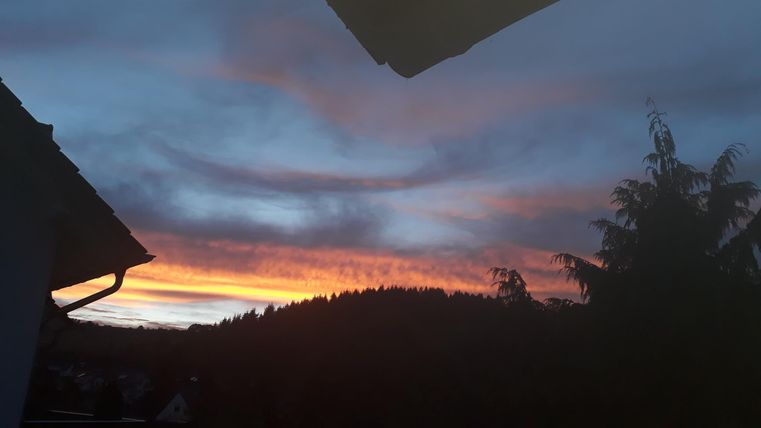 Un magnifique coucher de soleil derrière des montagnes avec un ciel orange et bleu. Les silhouettes d'une maison et d'arbres sont visibles au premier plan.