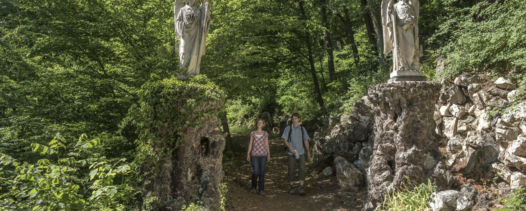 Zwei Personen spazieren auf einem Waldweg, flankiert von zwei Engelsstatuen auf steinernen Sockeln, umgeben von &uuml;ppigem Gr&uuml;n., &copy; TI Hocheifel-N&uuml;rburgring, Kappest