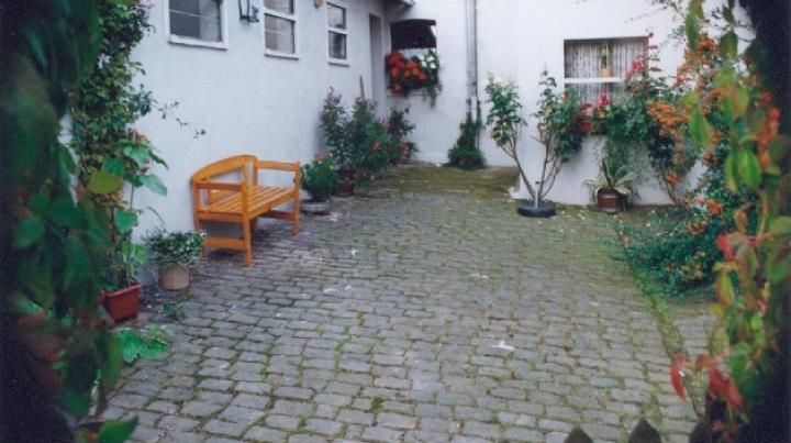 A small, paved courtyard with a wooden bench and blooming plants. The walls are white and some windows are visible.