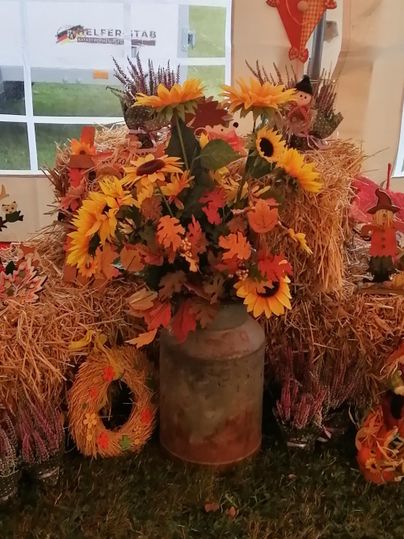 Eine herbstliche Dekoration mit Heuballen und bunten Blumen, darunter Sonnenblumen. Ein rustikales Gefäß ergänzt die Stimmung.