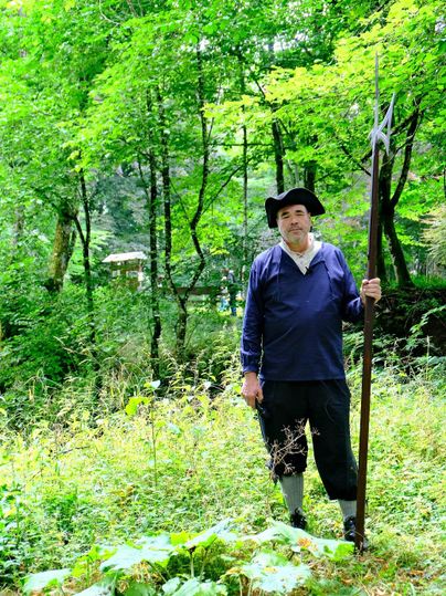 Ein Mann in historischer Kleidung steht in einem grünen Wald. Er hält einen langen Stock und schaut in die Kamera.