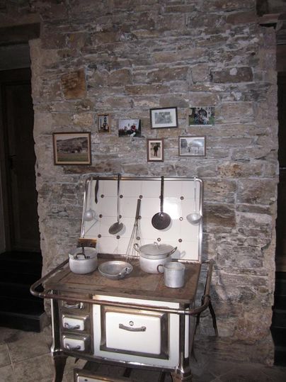 An old kitchen unit made of metal stands in front of a stone wall. Pictures in various frames hang on the wall.