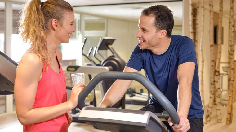 Twee personen in een sportschool praten met elkaar. Op de achtergrond zijn fitnessapparaten zichtbaar.