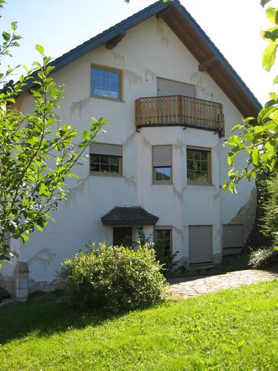 Une grande maison avec un balcon et un jardin. La façade est partiellement inachevée, entourée d'arbres et de buissons.