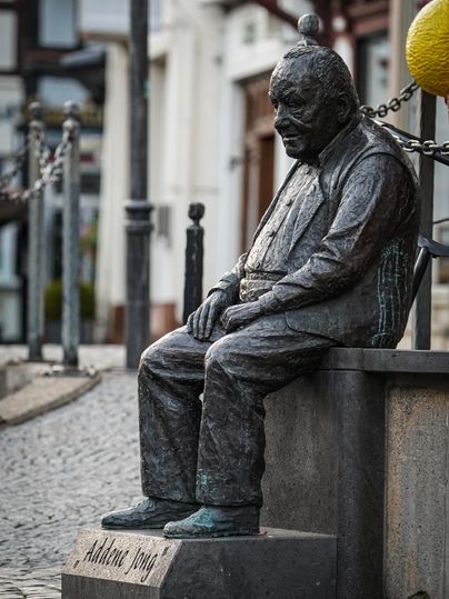 A bronze statue of a seated man on a stone bench. In the background, houses and a yellow sphere are visible.