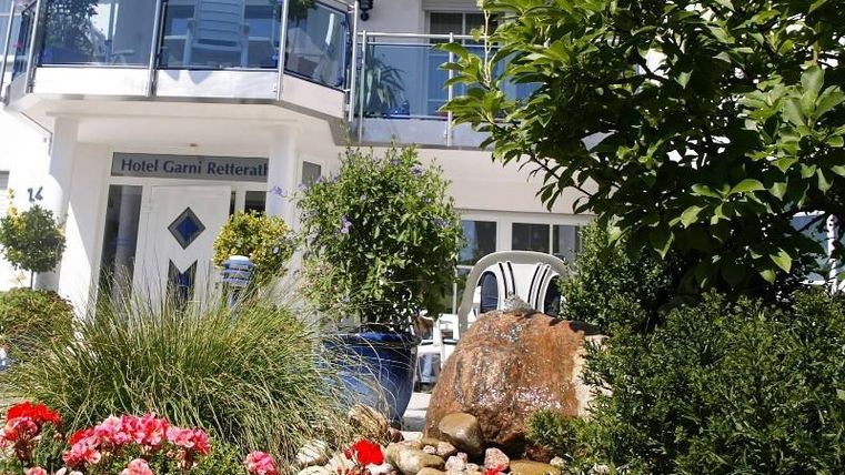 Ein hübsches Hotel mit einem gepflegten Garten und bunten Blumen. Der Eingang ist einladend mit einer Terrasse und grünen Pflanzen.