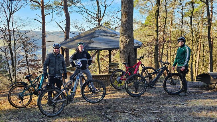 Vier Männer stehen mit ihren Mountainbikes in einem Waldgebiet. Im Hintergrund ist ein kleiner Pavillon und Bäume zu sehen.