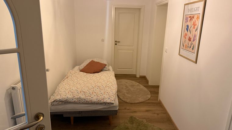 Een kleine ruimte met een eenpersoonsbed en een decoratief kussen. Aan de muren zijn een deur en een afbeelding met bloemenmotieven.