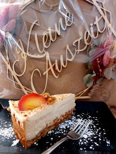 Un morceau de cheesecake sur une assiette noire avec du sucre glace. En arrière-plan, il y a une inscription décorative "Kleine Auszeit".