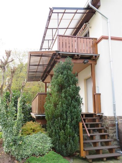 Une maison confortable avec un balcon en bois et un escalier. Le jardin est vert avec une variété de plantes.