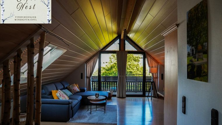Een gezellige woonkamer met een grijze bank en een ronde tafel. Grote ramen bieden een mooi uitzicht op de groene omgeving.