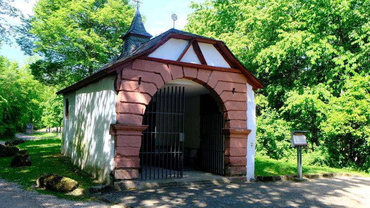 Een kleine kapel met een decoratieve ingang en een spitse dak. Ze staat omringd door groene bomen in een rustige omgeving.
