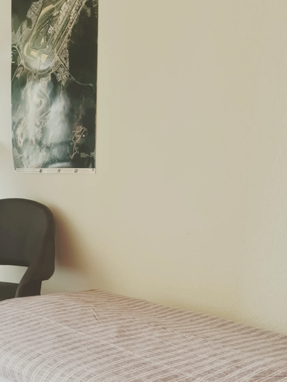 A simple room with a chair and a blanket. On the wall hangs a poster of an airport.