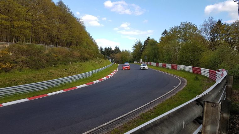 Eine Rennstrecke mit zwei Autos, die um eine Kurve fahren. Umgeben von grünen Bäumen und einem blauen Himmel.