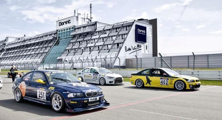 Een groep racewagens rijdt op een racecircuit. Op de achtergrond is het gebouw van het racecentrum te zien.