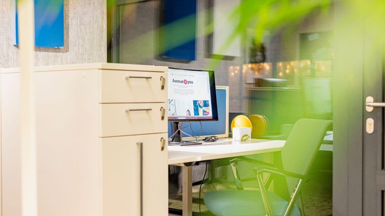 Ein modernes Büro mit einem Schreibtisch, einem Computer und einem Stuhl. Im Vordergrund ist ein Aktenschrank und etwas Pflanzenbewuchs zu sehen.