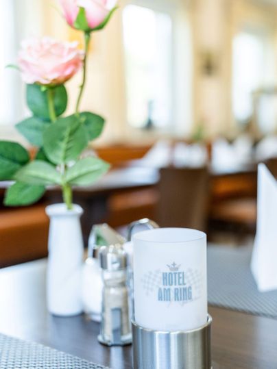 Een tafel in een restaurant met een vaas die een roze bloem heeft. Naast de vaas staat een kaarsenhouder met de tekst "HOTEL AM RING".