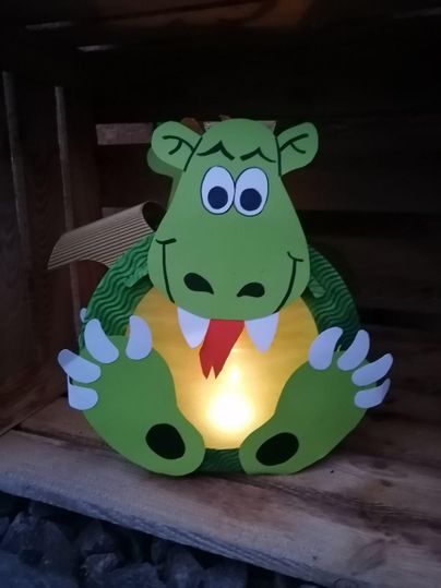 Eine bunte Drachenlampe mit großen, freundlichen Augen und einem lächelnden Gesicht. Sie steht in einem Holzregal und leuchtet sanft.