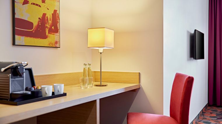 A cozy hotel room area with a table, a lamp, and a red chair. A picture hangs on the wall and a television is installed.
