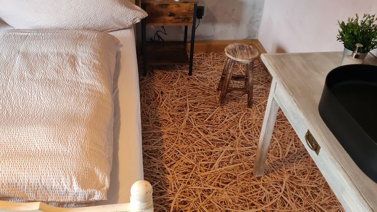 Een gezellige kamer met een bed, een tafel en een kruk. De vloer is bedekt met stro materiaal en de muren zijn geschilderd in zachte kleuren.