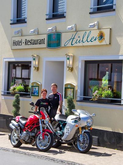 A motorcycle rider couple stands in front of the Hotel-Restaurant Hüller. Next to them are two motorcycles, one red and one silver.