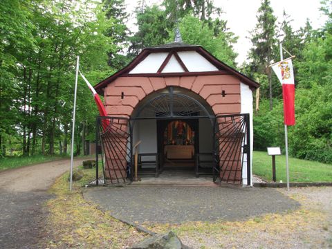 A small pavilion in the woods with an open entrance door. Flags hang on both sides and the path is surrounded by trees.