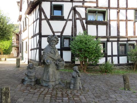 A statue of a woman with two children stands on the pavement in front of a half-timbered house. The surroundings are green and inviting.