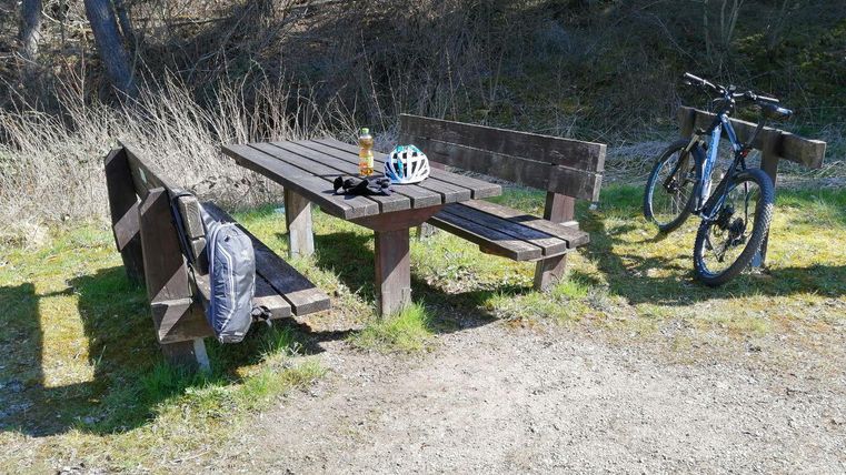 Ein Holz-Tisch mit Bänken in der Natur, neben einem Fahrrad. Auf dem Tisch liegen eine Wasserflasche und ein Fahrradhelm.