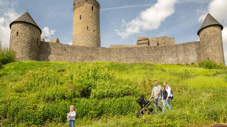 Eine Familie mit einem Kinderwagen steht auf einem Weg vor einer alten Burg. Im Hintergrund sieht man die Burgmauern und Türme, umgeben von grünen Wiesen.