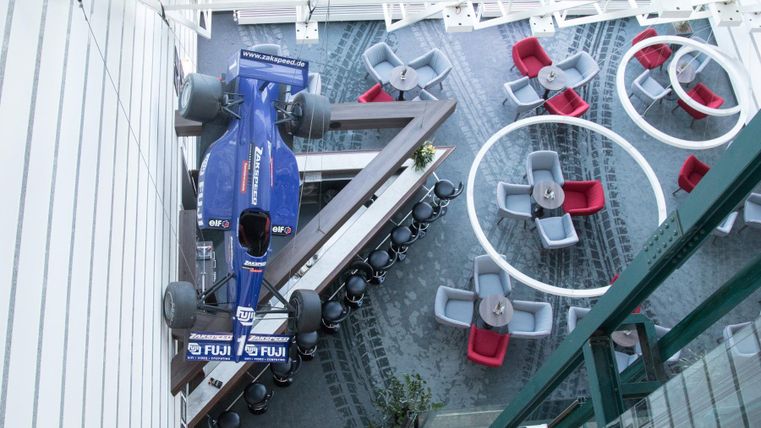 A Formula 1 car hangs over a modern, stylish restaurant area. The decor consists of gray and red chairs as well as round tables.