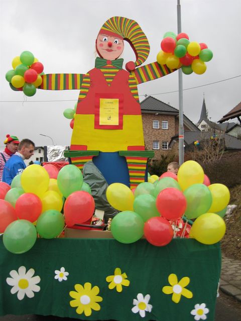Een kleurrijke carnavalswagen met een grote kartonnen figuur van een clownachtige fabrikant en vele ballonnen. De scene is feestelijk en vrolijk ingericht.