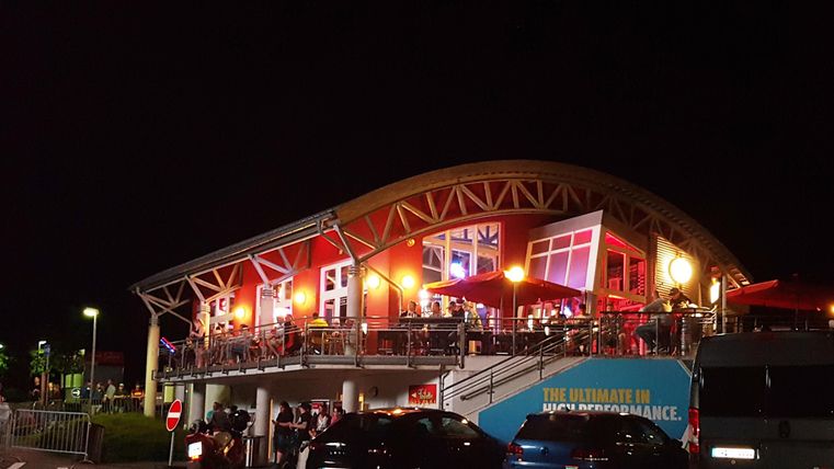 Un bâtiment de restaurant moderne la nuit, illuminé par des lumières colorées. Des terrasses décorées invitent à rester.