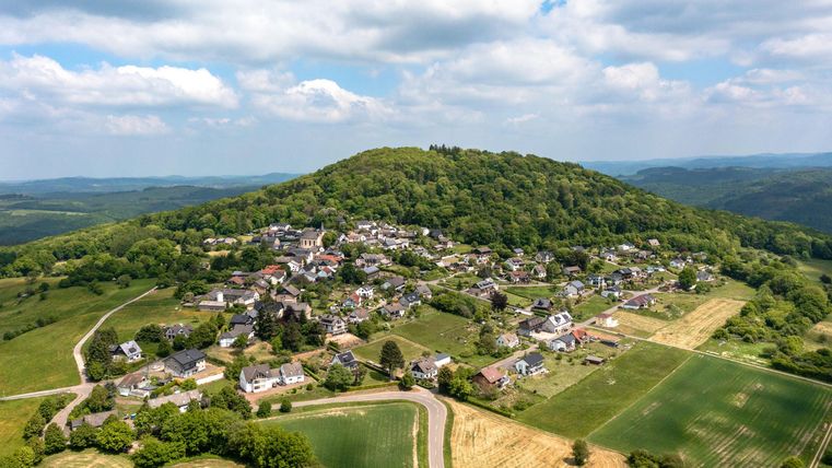 Eine malerische Landschaft mit einem kleinen Dorf am Fuße eines bewaldeten Hügels. Im Hintergrund sind grüne Felder und sanfte Hügel sichtbar.