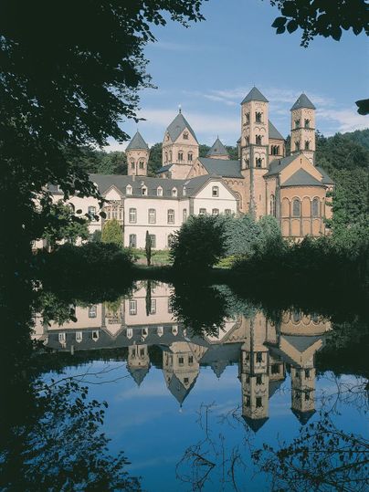 Ein beeindruckendes Gebäude mit Türmen spiegelt sich im ruhigen Wasser eines Sees. Umgeben von Bäumen und einer malerischen Landschaft.