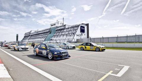 A group of racing cars is driving on a racetrack. In the background, the building of the racing center can be seen.