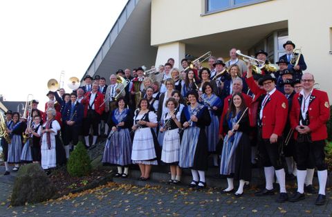 Een grote groep mensen staat in traditionele klederdracht voor een gebouw. Velen hebben muziekinstrumenten bij zich en de sfeer is feestelijk.