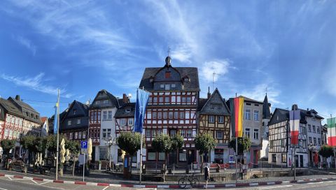 A beautiful historic town square with several half-timbered houses. The sky is clear and blue, and flags are fluttering on the buildings.