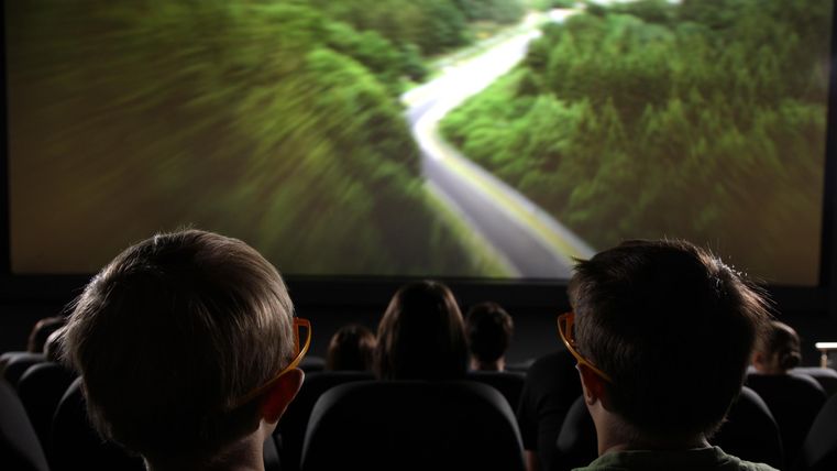 Zwei Kinder sitzen im Kino und schauen sich einen Film an. Auf der Leinwand ist eine landschaftliche Aufnahme mit einer kurvenreichen Straße zwischen Bäumen zu sehen.